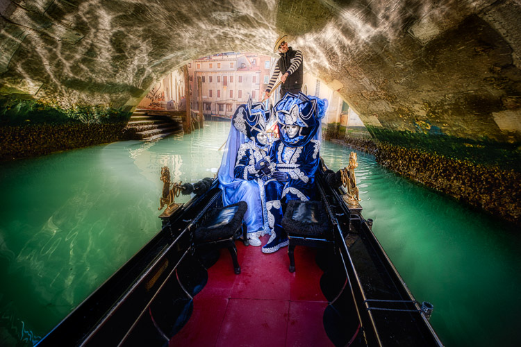 Costumed and masked characters in gondola on Venice canal passing under a bridge with reflection on underside of bridge